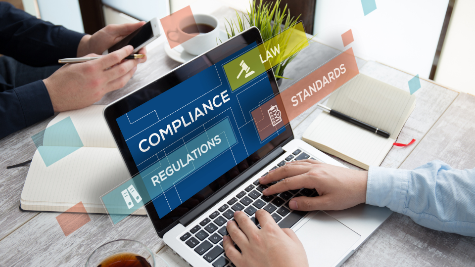 Alt text: A laptop on a desk with the words "Compliance," "Law," "Regulations," and "Standards" floating above the screen in bold text