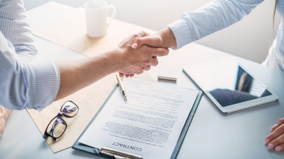 Alt text: Two people shaking hands over an employment contract placed on a desk