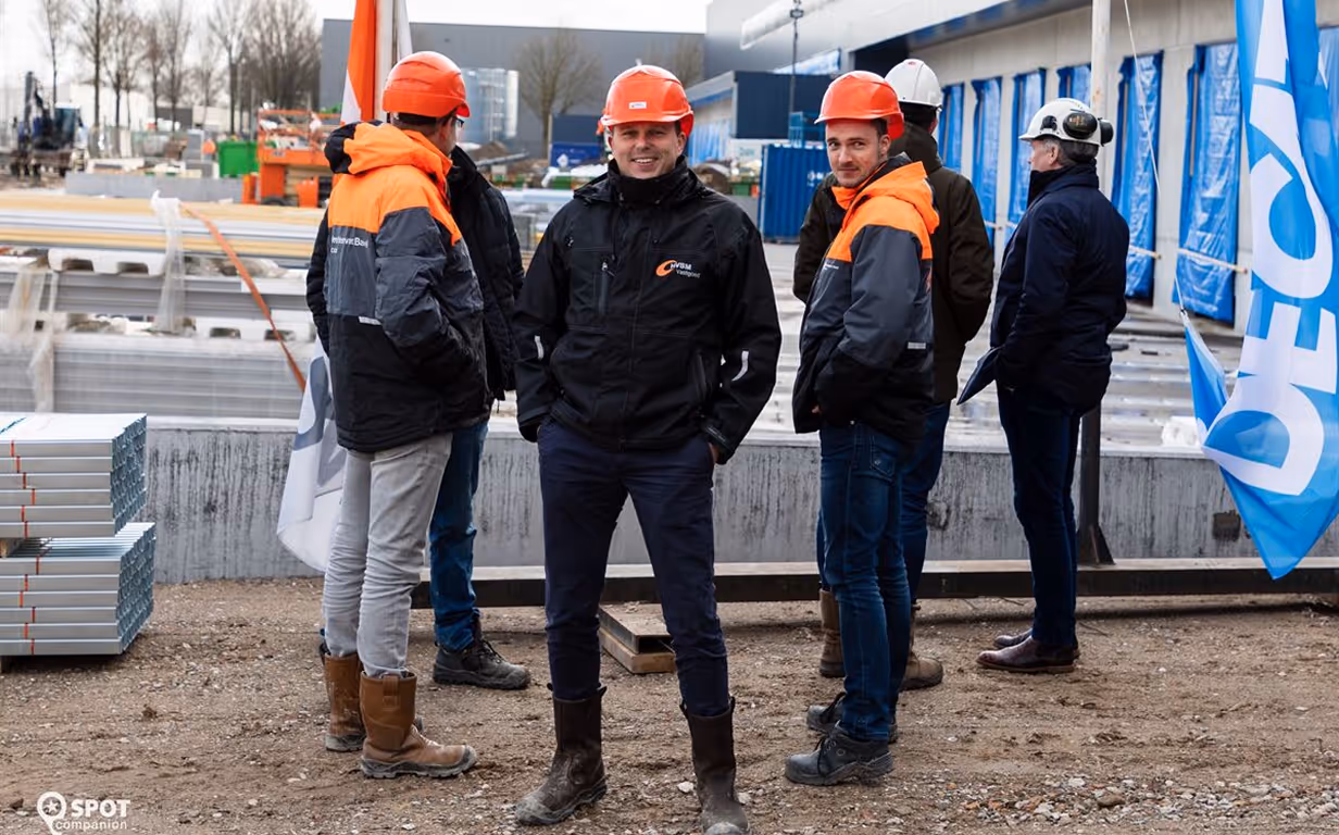 Group of construction workers in helmets and jackets standing at a building site with materials nearby.