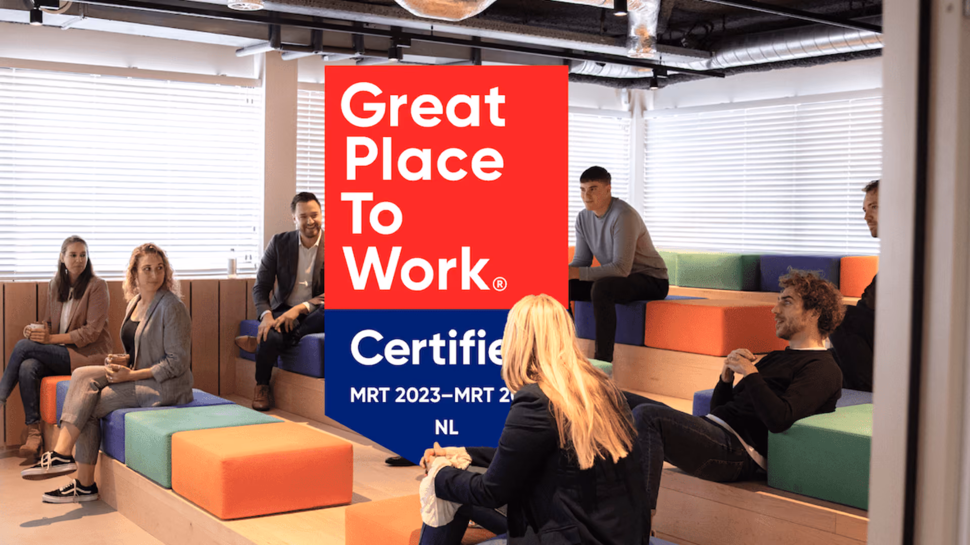 Group of six people sitting on colorful steps in a modern office with 'Great Place To Work Certified' sign.