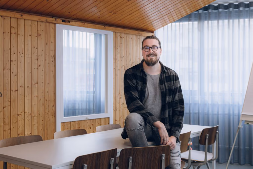 Smiling man with glasses and beard sitting on table in wood-paneled room with chairs and window.