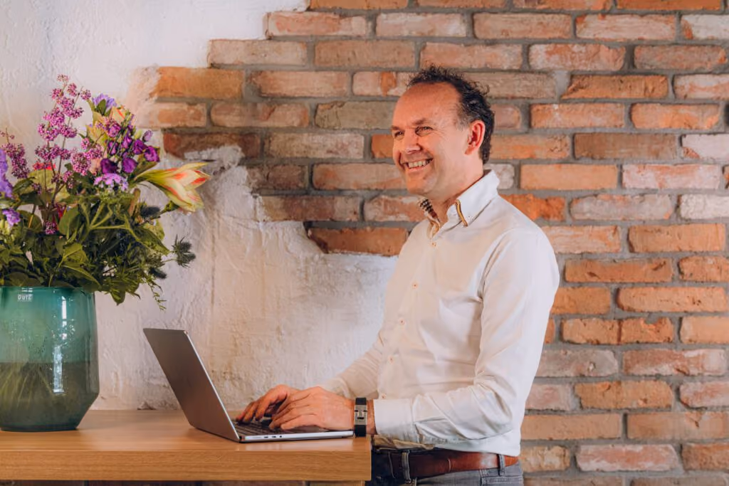 Smiling man in white shirt typing on a laptop at a wooden desk with a green vase of flowers.