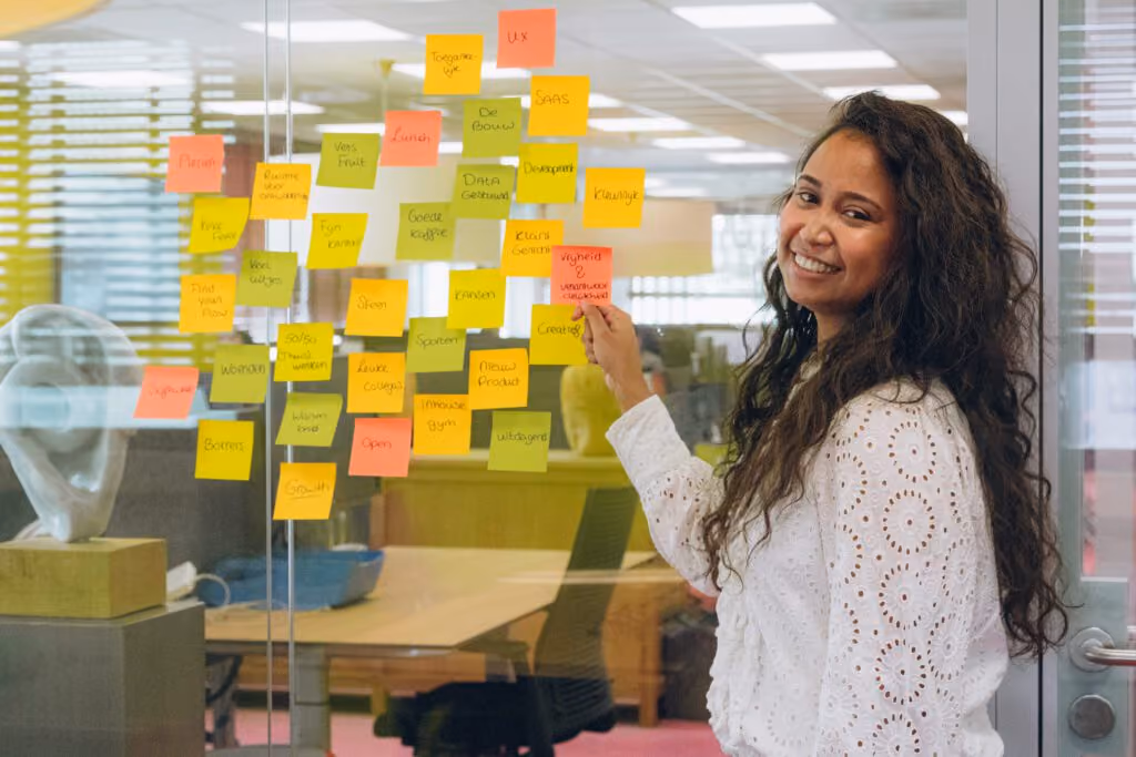 Smiling woman with long curly hair placing a sticky note on a glass wall covered with colorful notes.
