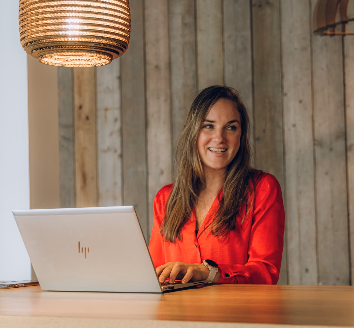Woman in red shirt sitting at wooden desk with laptop, smiling and looking to the side.