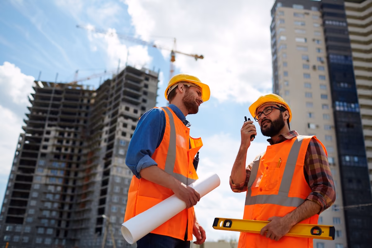 Two construction workers in helmets and vests discuss plans at a building site, one holding blueprints.