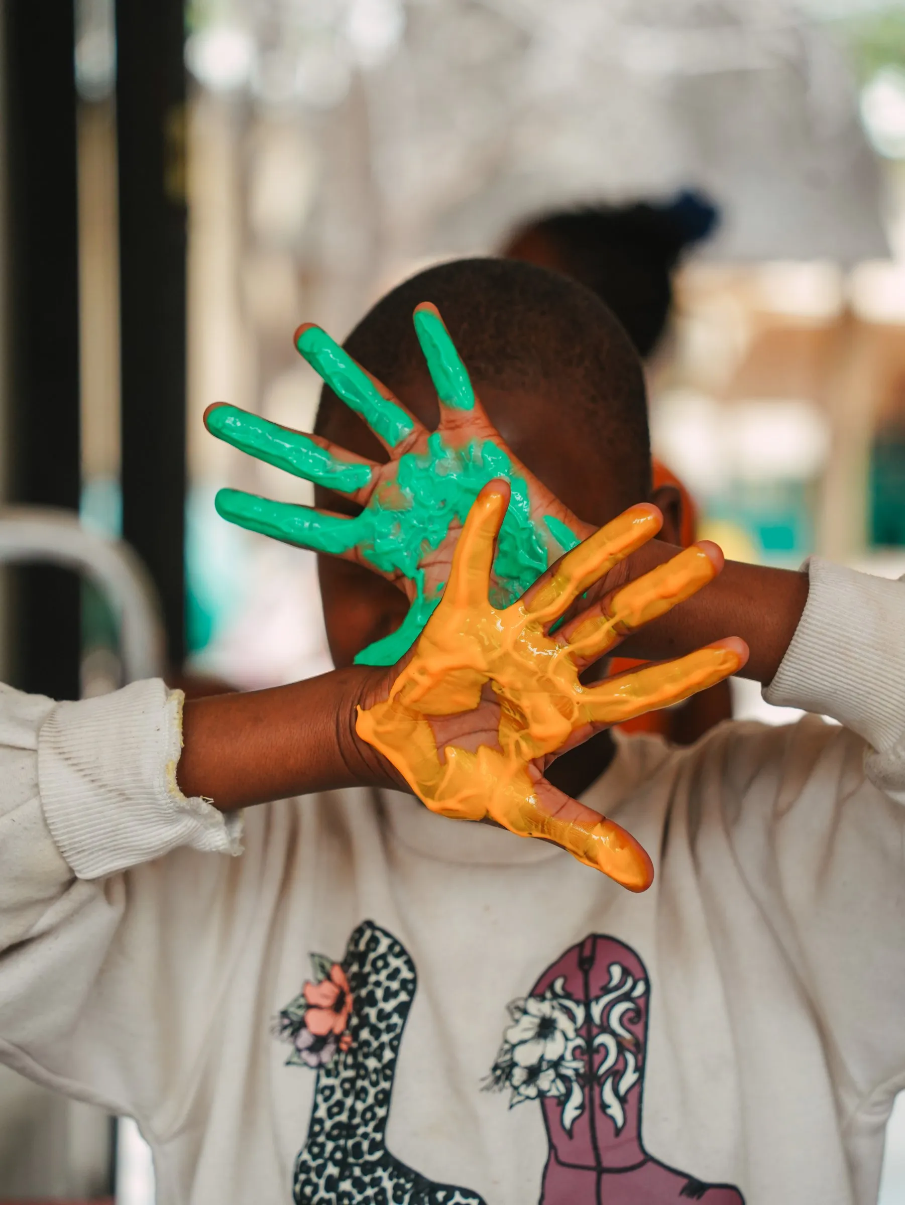 A child at Lorato House holding up two hands covered in green and orange paint