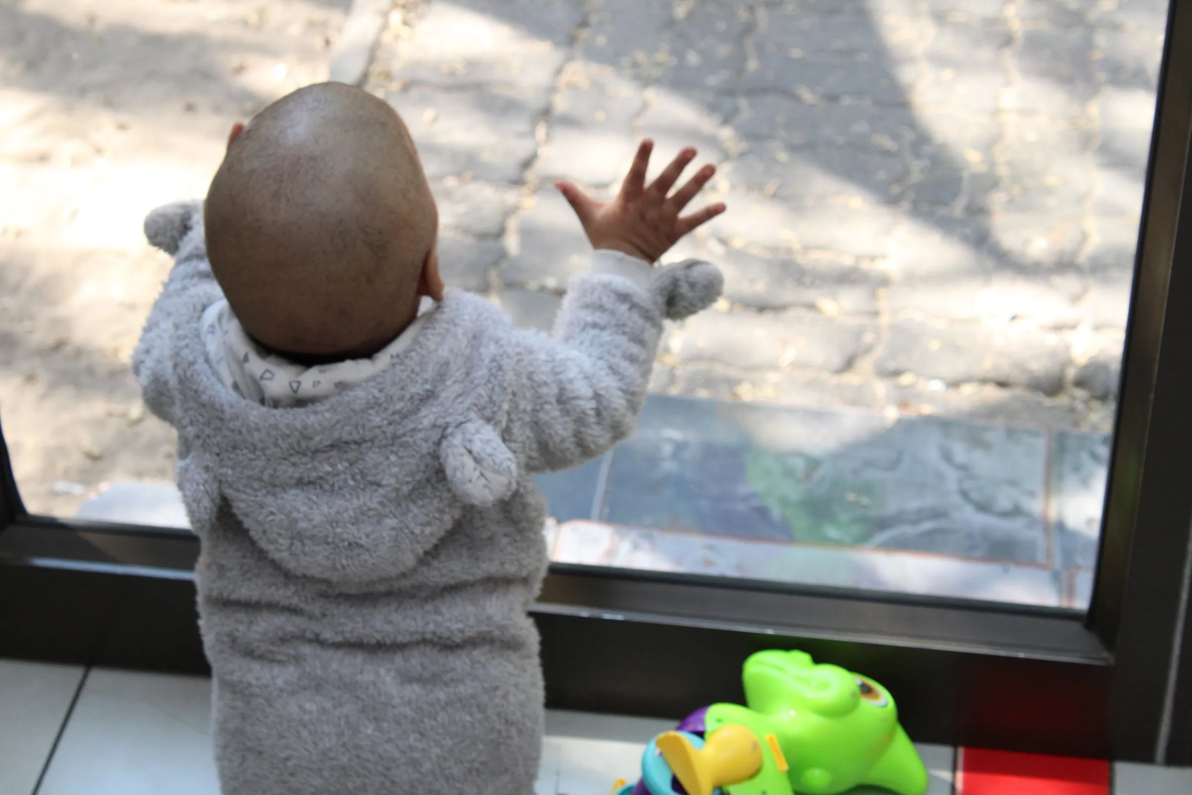 A baby in a soft grey onesie reaching up to a window at Lorato House