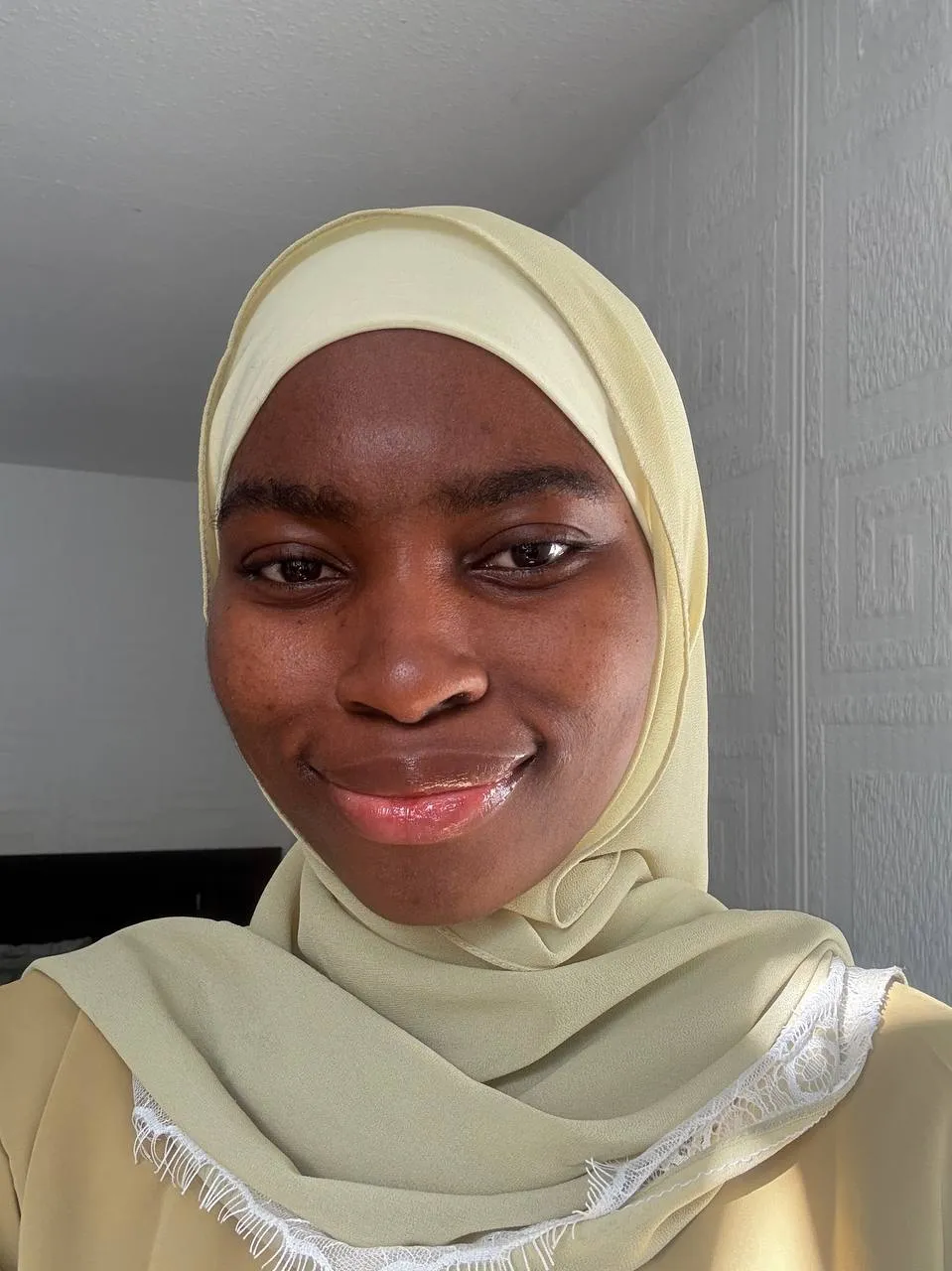 Close-up of a woman smiling, wearing a light yellow hijab and matching outfit, indoors near textured wall.