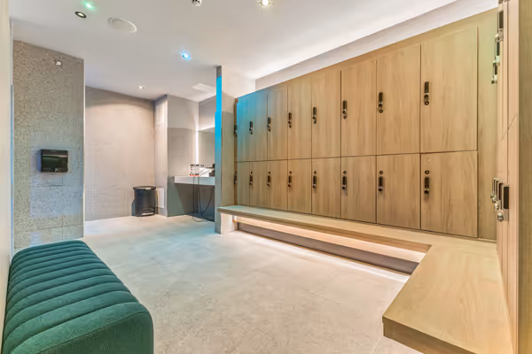 Modern locker room with wooden lockers, glass shower area, green bench, and beige tiled floor.