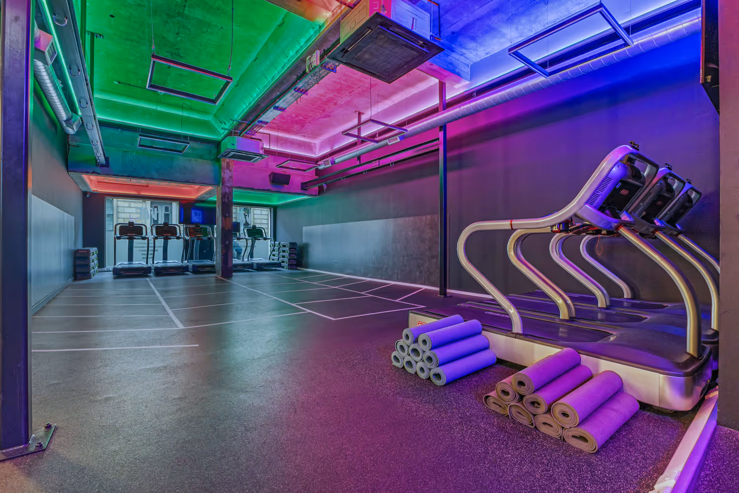 Modern gym room with treadmills, colorful ceiling lights, and stacked exercise mats on the floor.