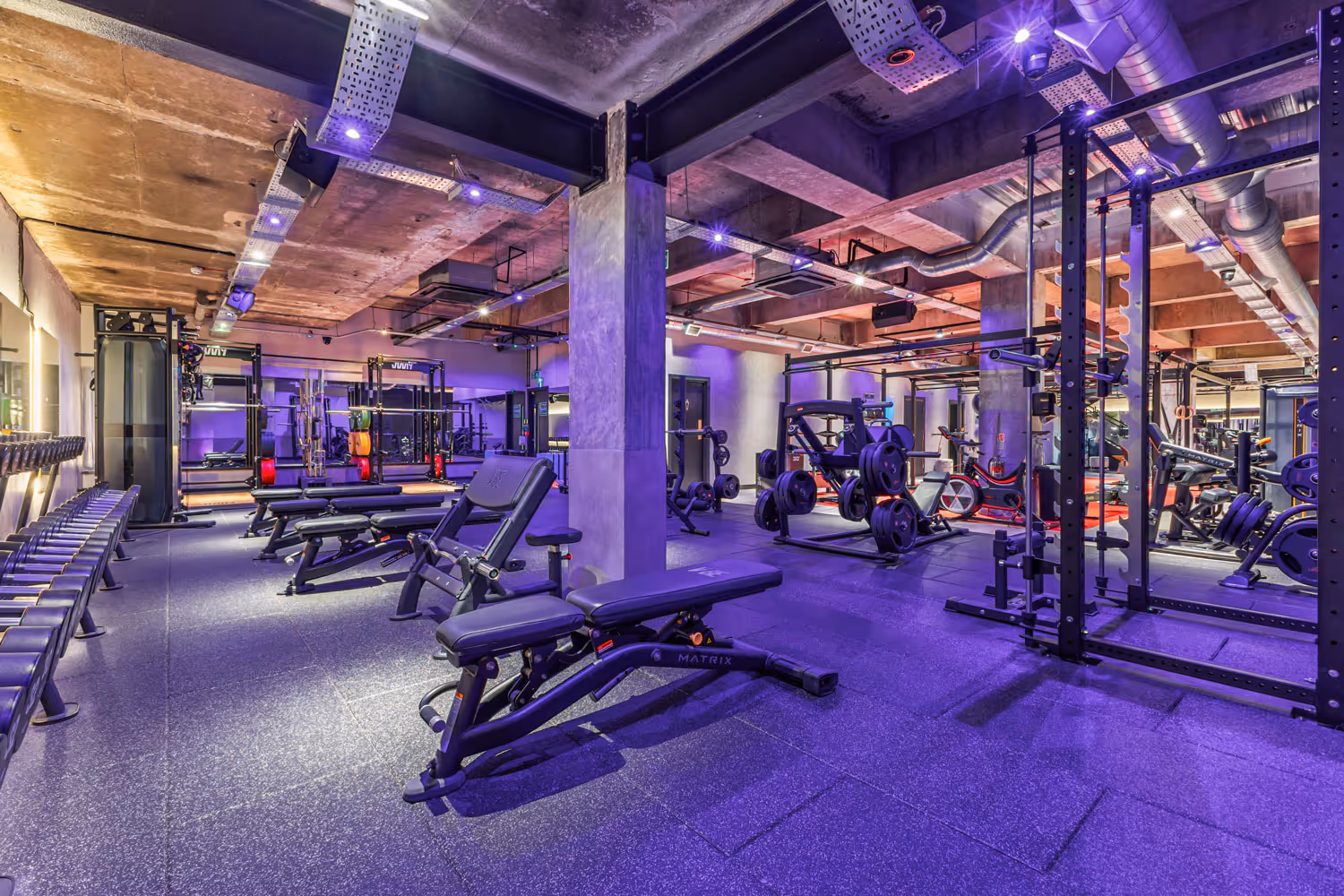 Modern gym weight area with benches, dumbbells, weight racks, and purple ambient lighting.