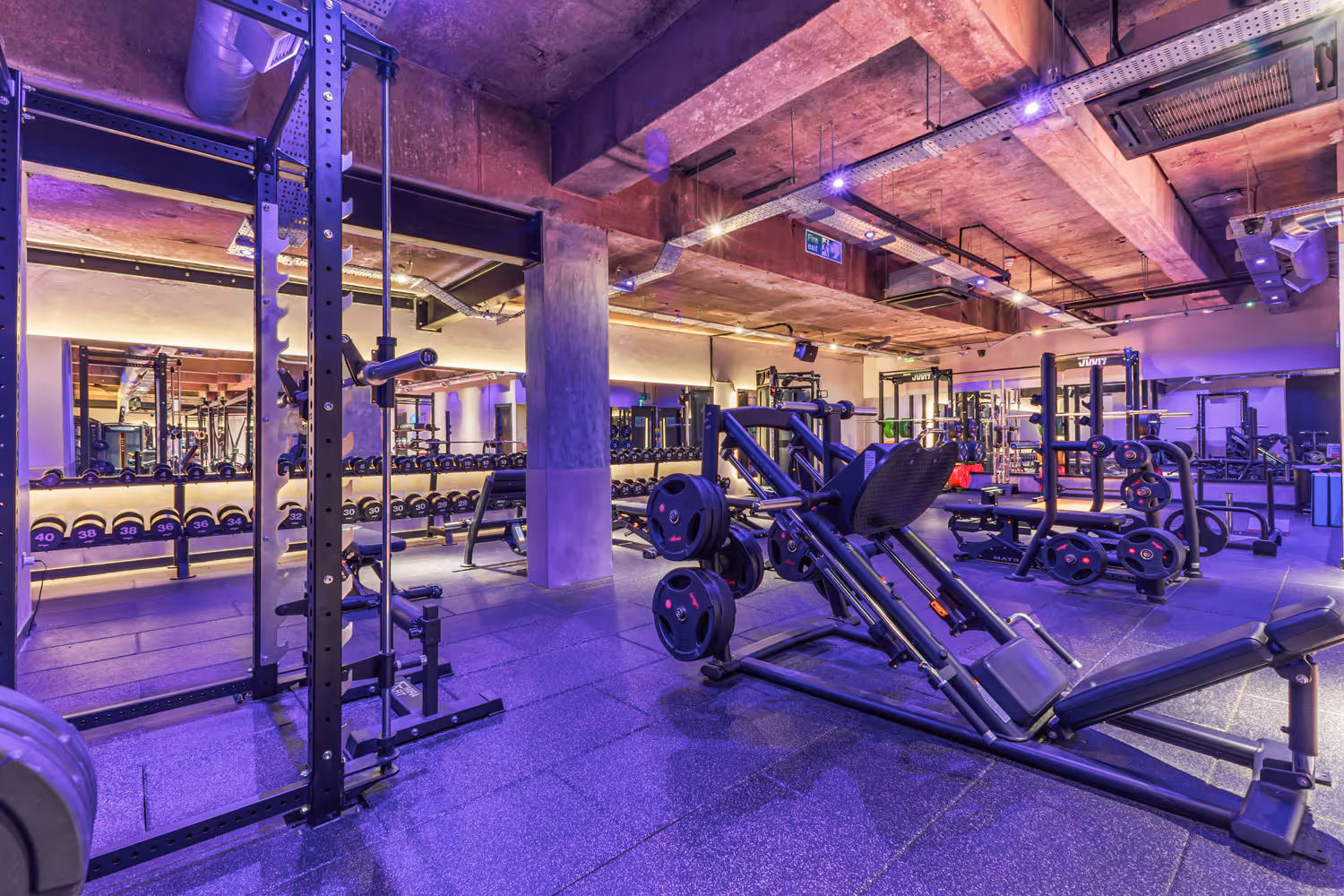 Well-lit gym area with weightlifting machines, racks of dumbbells, and exercise benches.