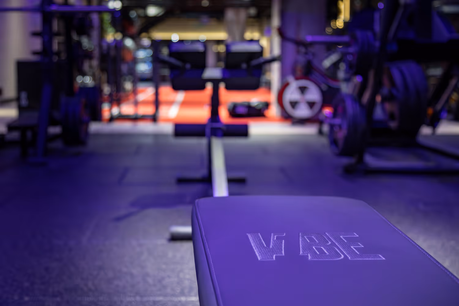 Close-up of a purple workout bench in a gym with blurred weights and machines in the background.