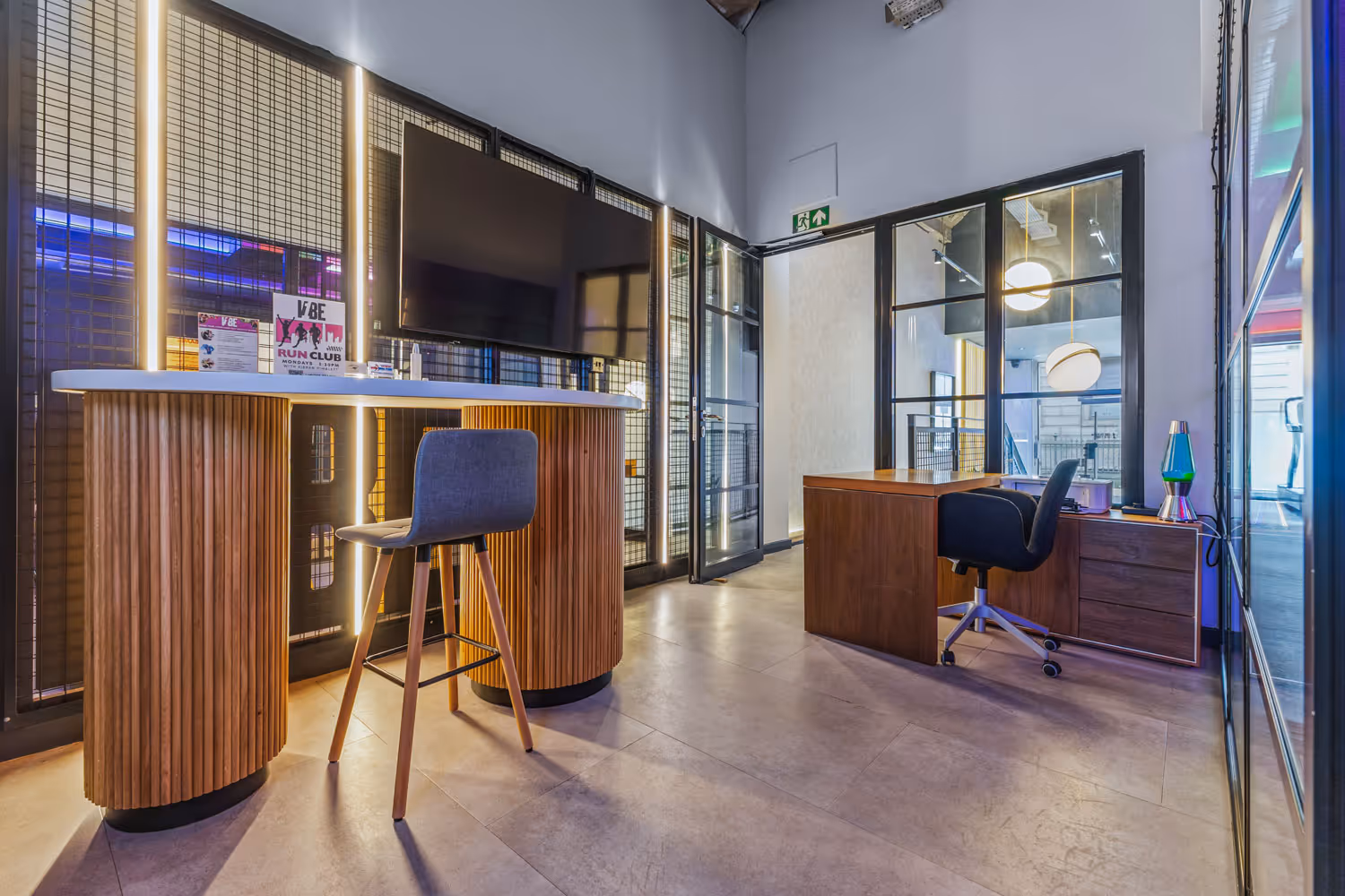 Modern office reception area with a wooden bar desk, stool, TV, and a workspace with a swivel chair and lava lamp.