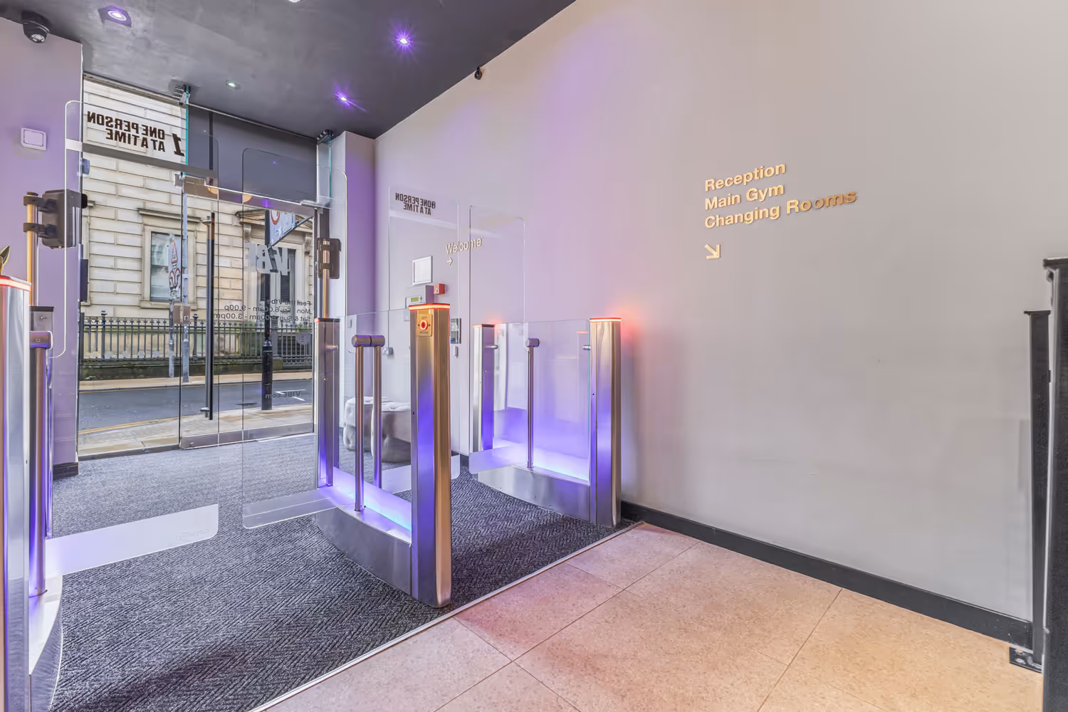 Entrance lobby with glass security gates and wall signage directing to reception, main gym, and changing rooms.