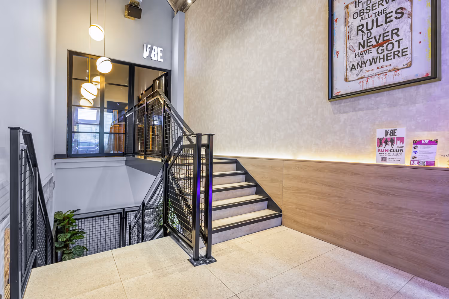 Modern staircase with black railing, hanging round lights, framed quote, and event flyers on wood-paneled wall.