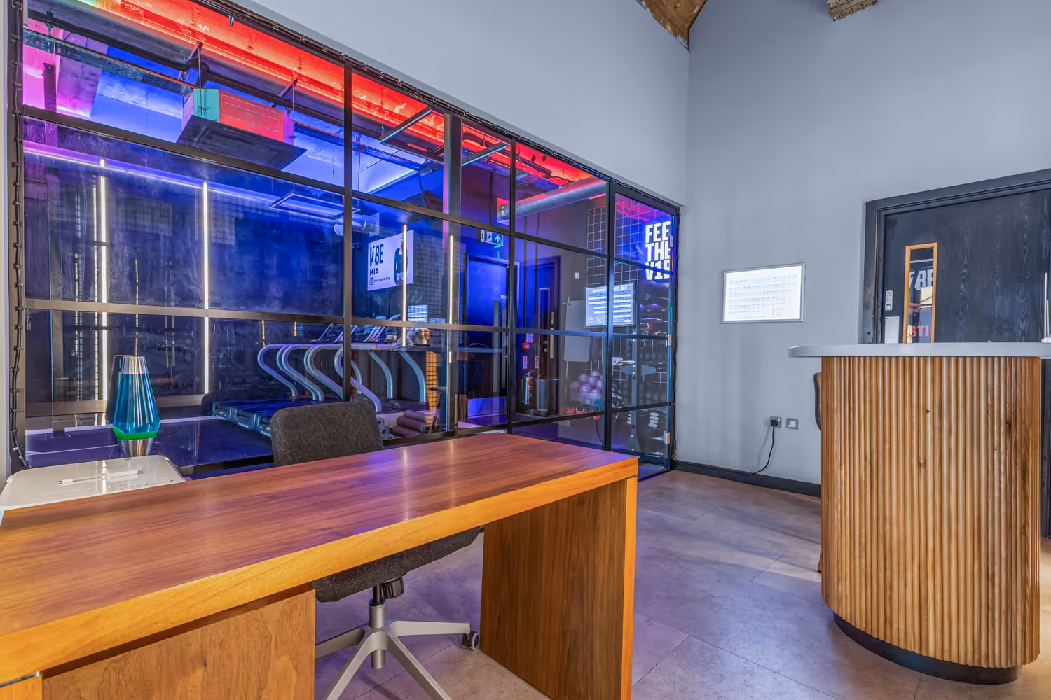 Modern gym reception with wooden desk and glass wall revealing treadmills and neon lighting inside gym area.