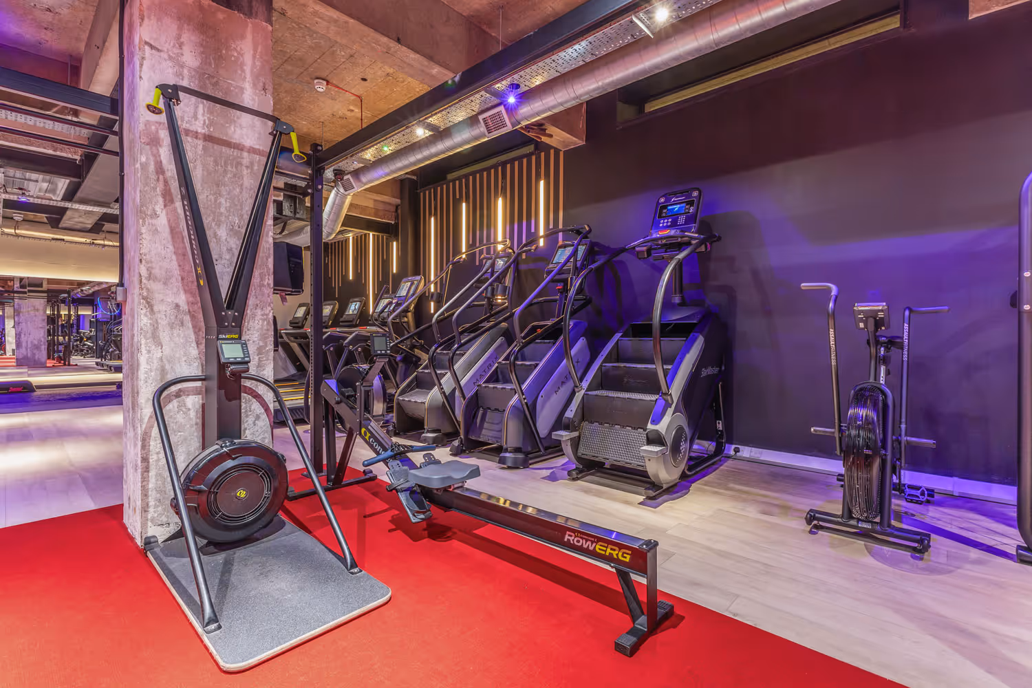 Interior of gym with cardio machines on a red and wood floor under industrial ceiling.