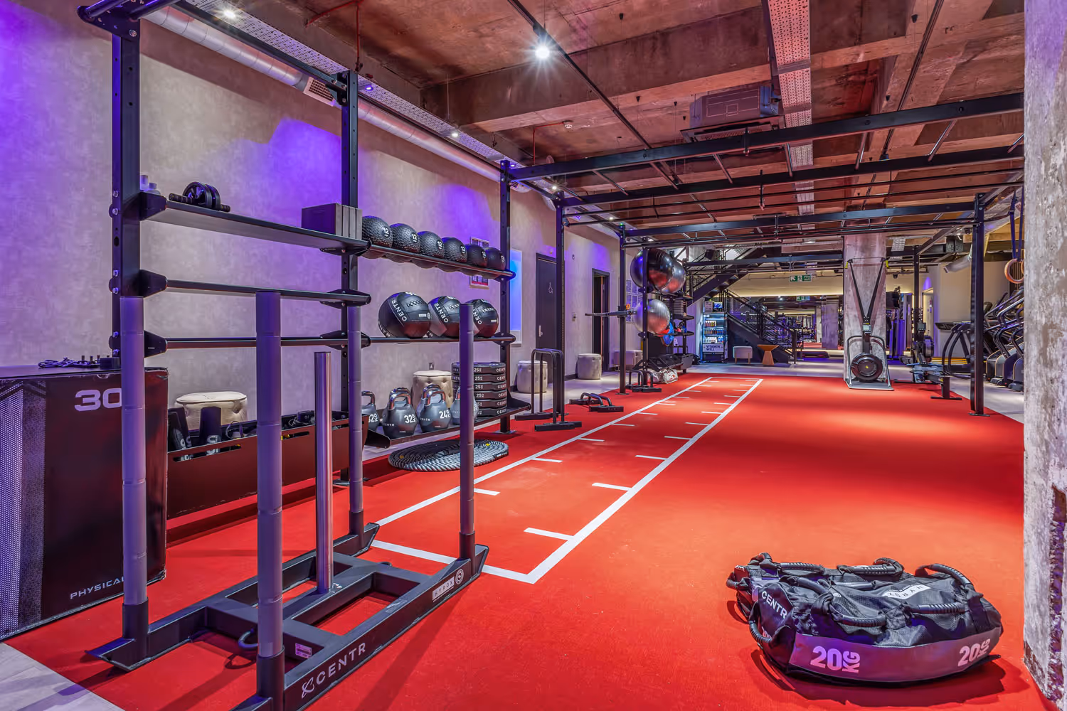 Gym area with red floor, weight sled, kettlebells, medicine balls, and fitness equipment under industrial ceiling.
