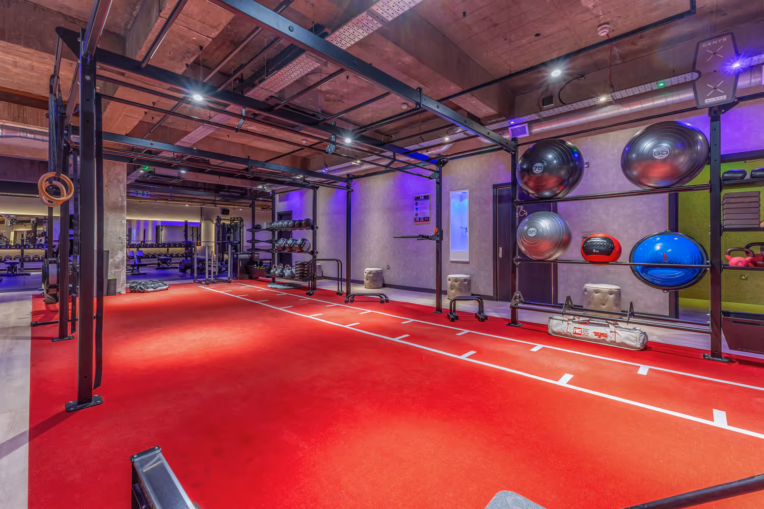 Modern gym with red flooring, exercise balls, kettlebells, rings, and weight racks under industrial ceiling.