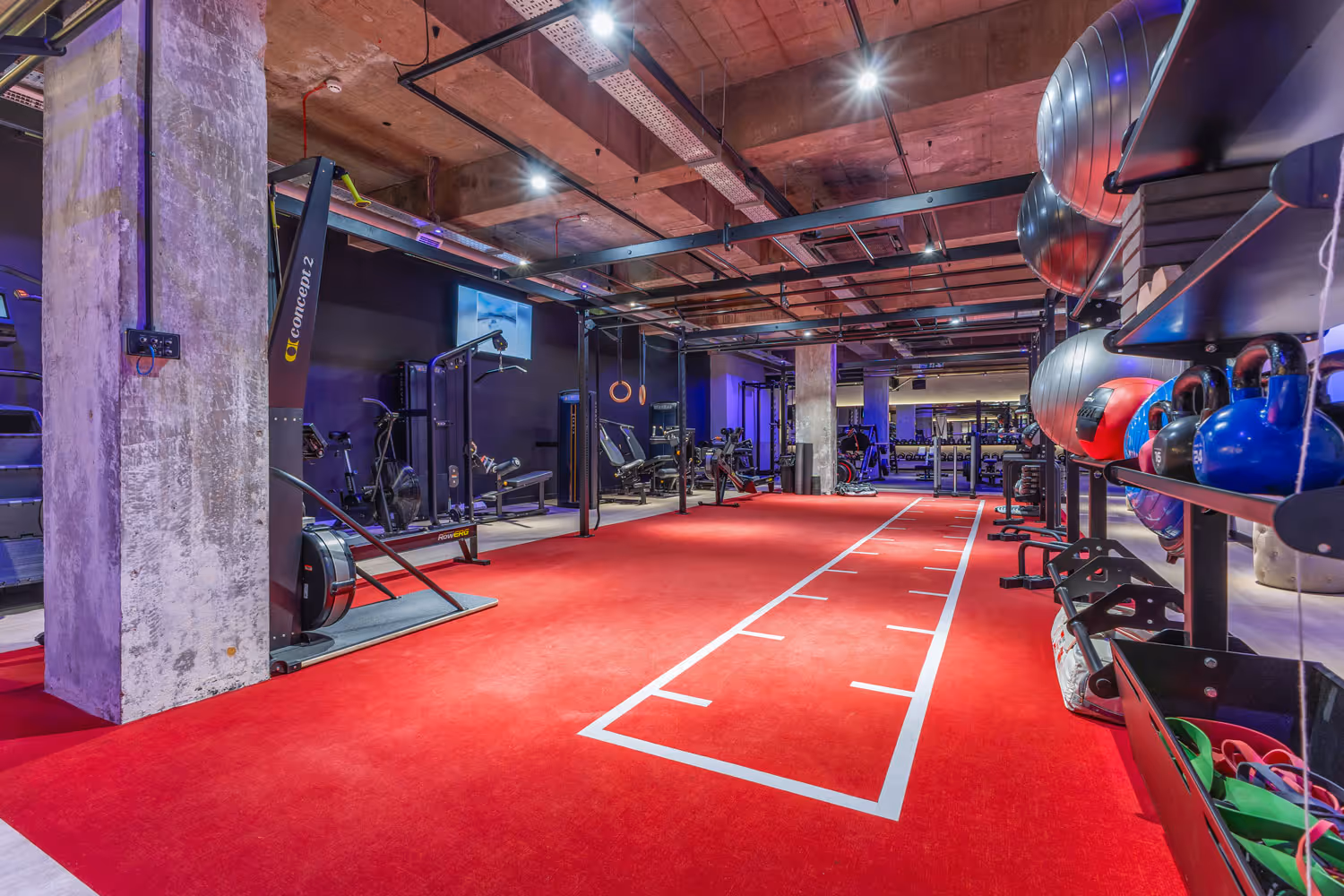 Modern gym with red workout turf, kettlebells, exercise balls, machines, and gymnastic rings.