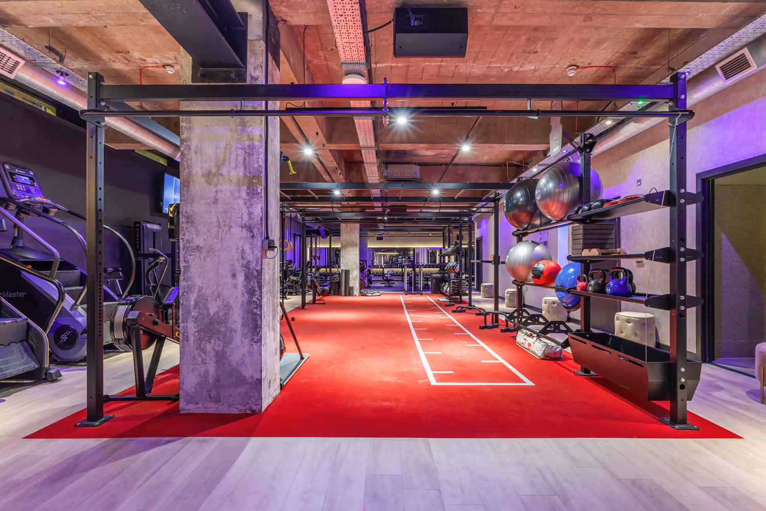Modern gym with red workout area, exercise balls, kettlebells, machines, and wooden flooring.