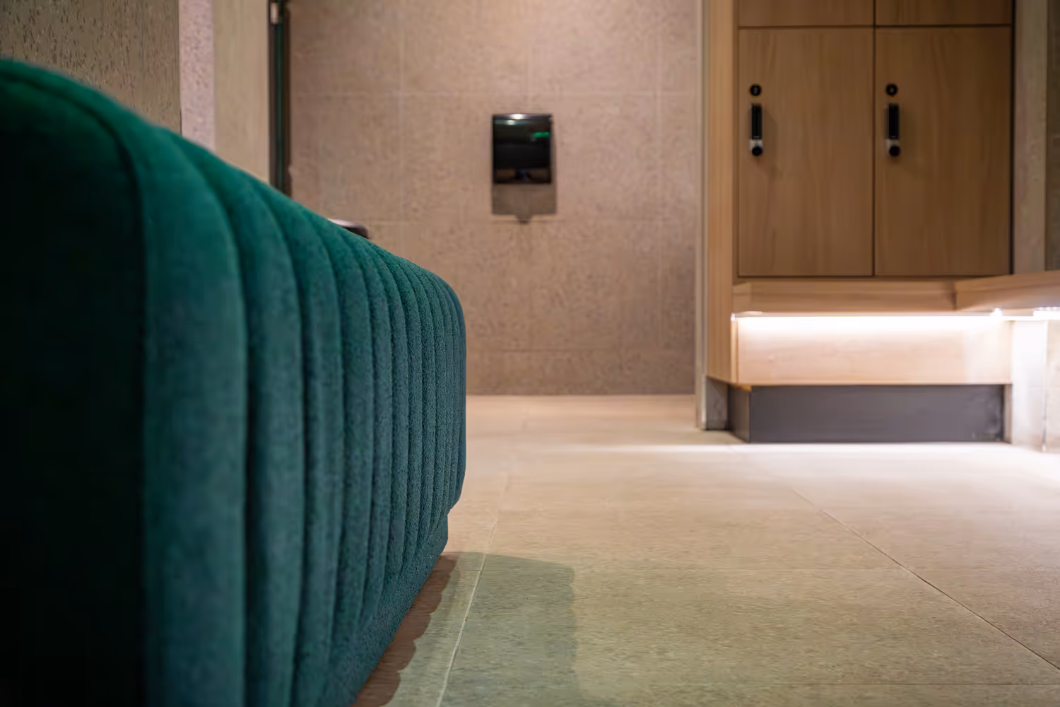 Green cushioned bench next to wooden lockers in a modern changing room with tiled floor and walls.