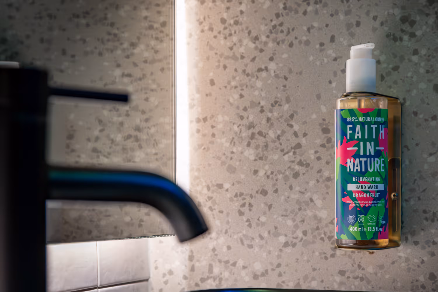 Wall-mounted Faith In Nature dragon fruit hand wash bottle beside a black faucet in a tiled bathroom.