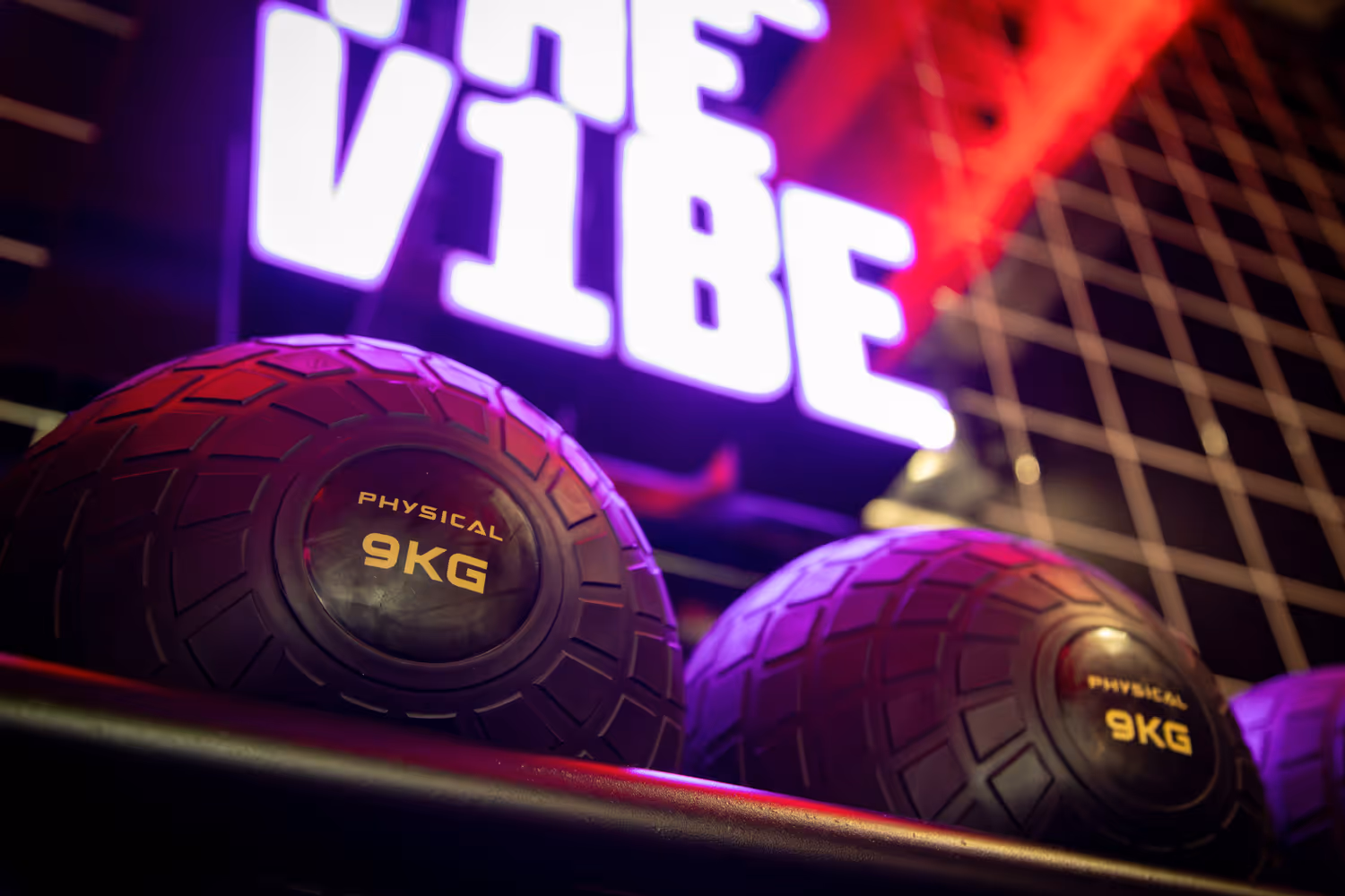 Two 9kg physical medicine balls on a rack with a blurred neon sign reading 'The Vibe' behind.