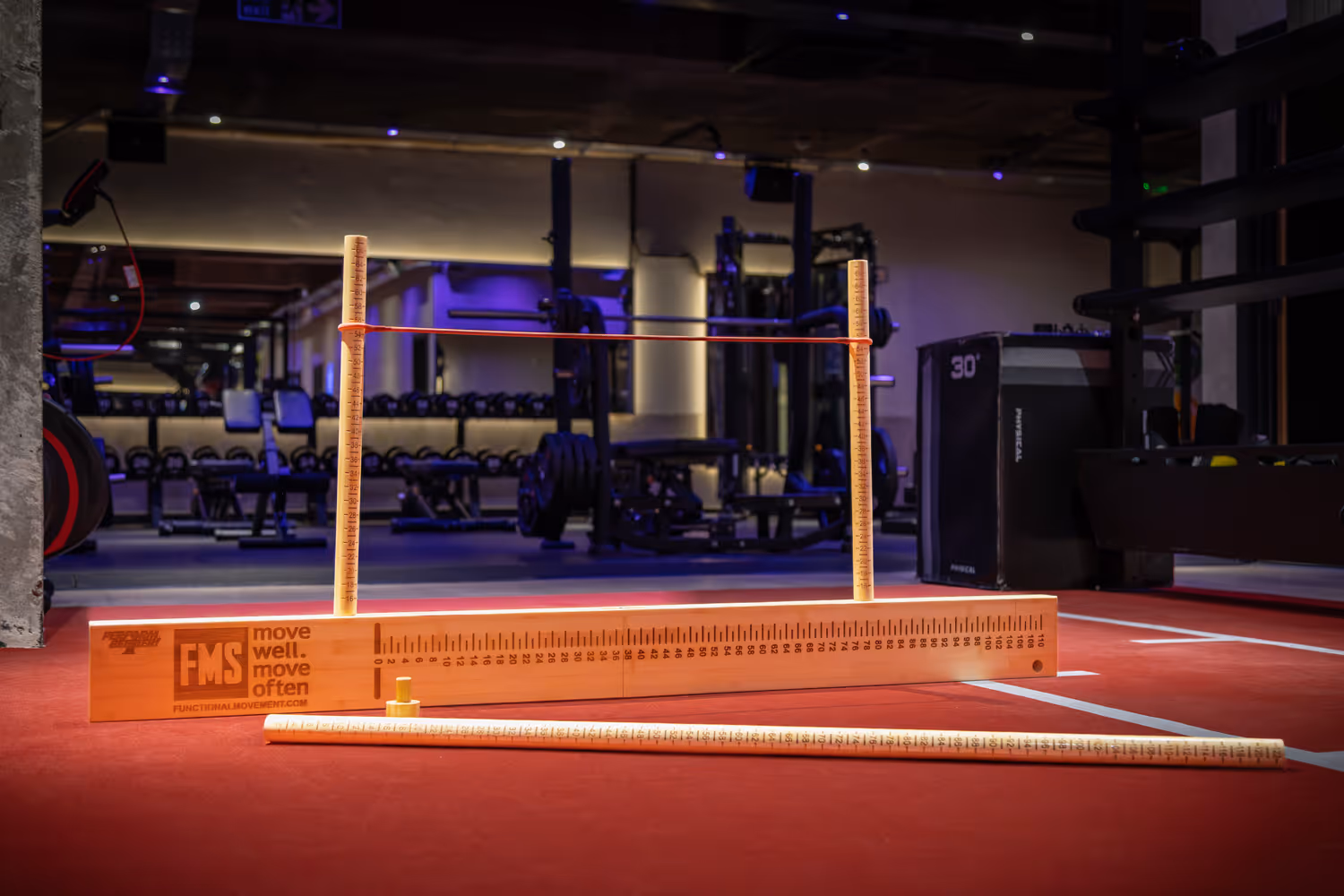 Functional Movement Screen tool with wooden measuring sticks and red rubber band on red gym floor.