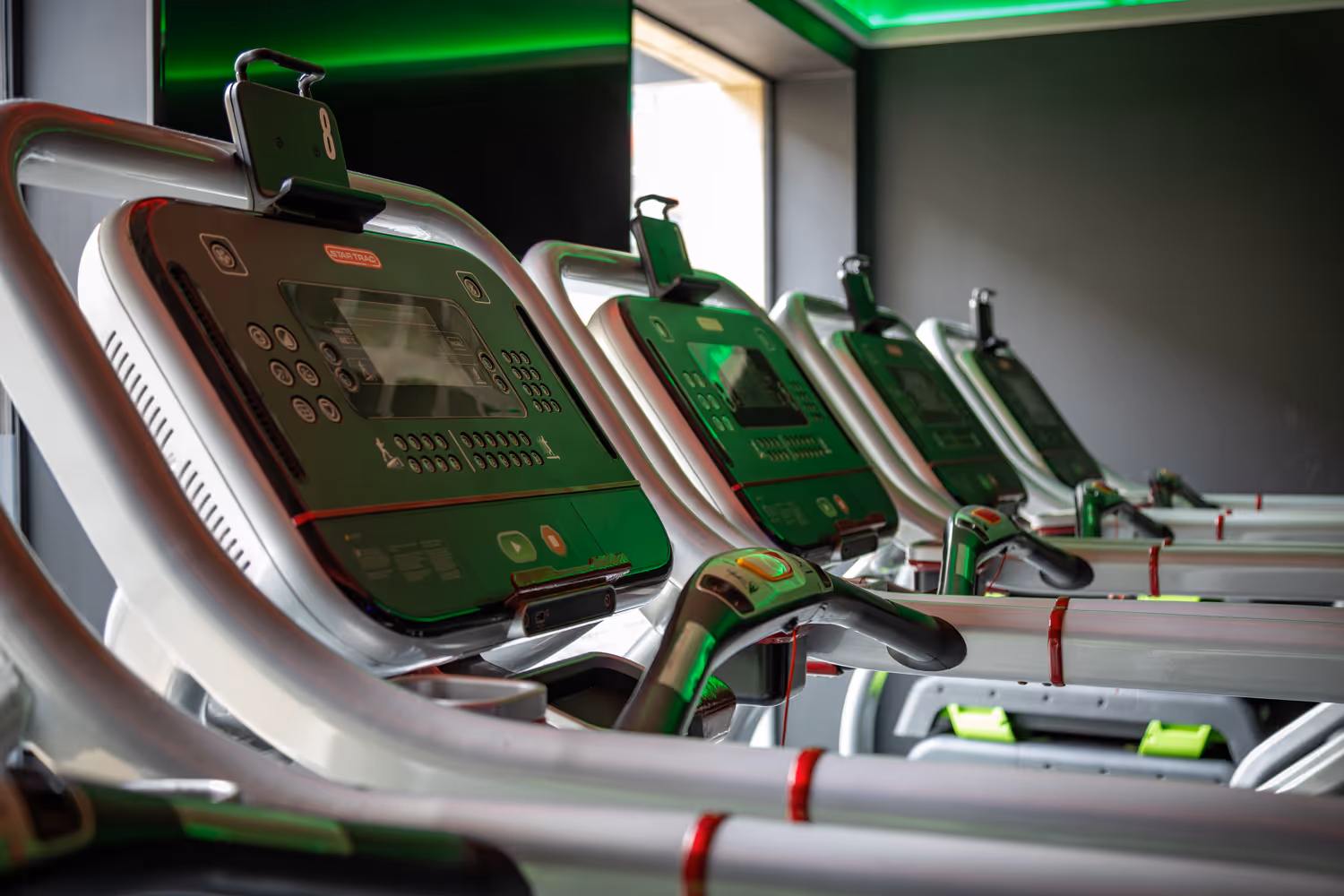 Close-up of a row of treadmills with digital display panels in a gym under green ceiling lights.