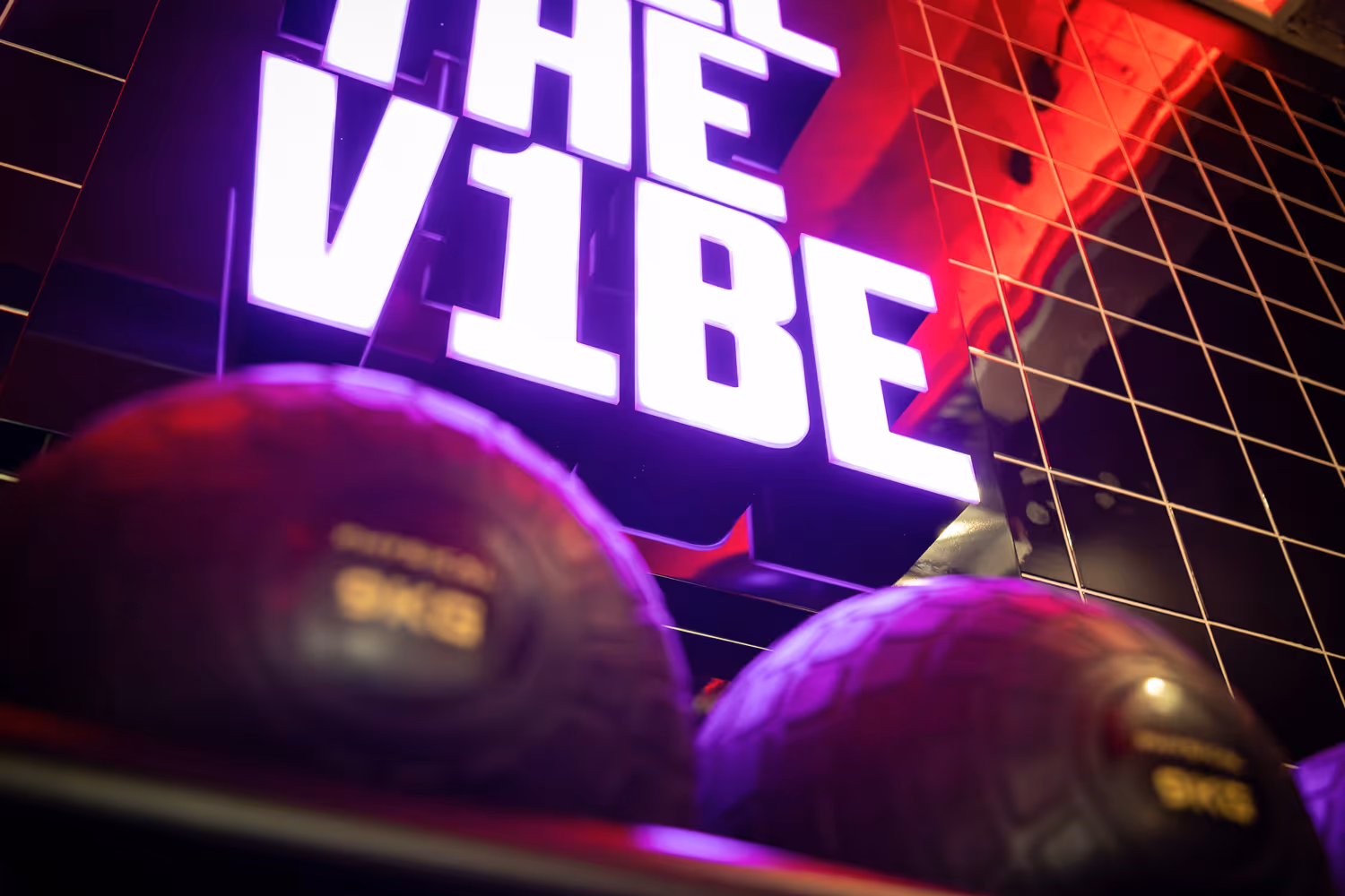 Close-up of medicine balls below a neon sign reading THE VIBE on a tiled wall.
