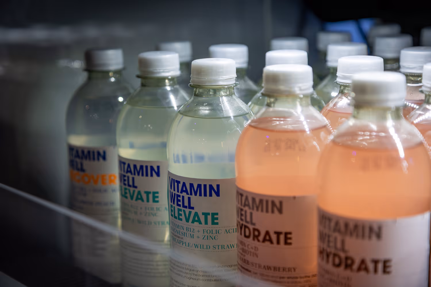 Rows of Vitamin Well bottled drinks in clear plastic bottles with white caps inside a fridge.
