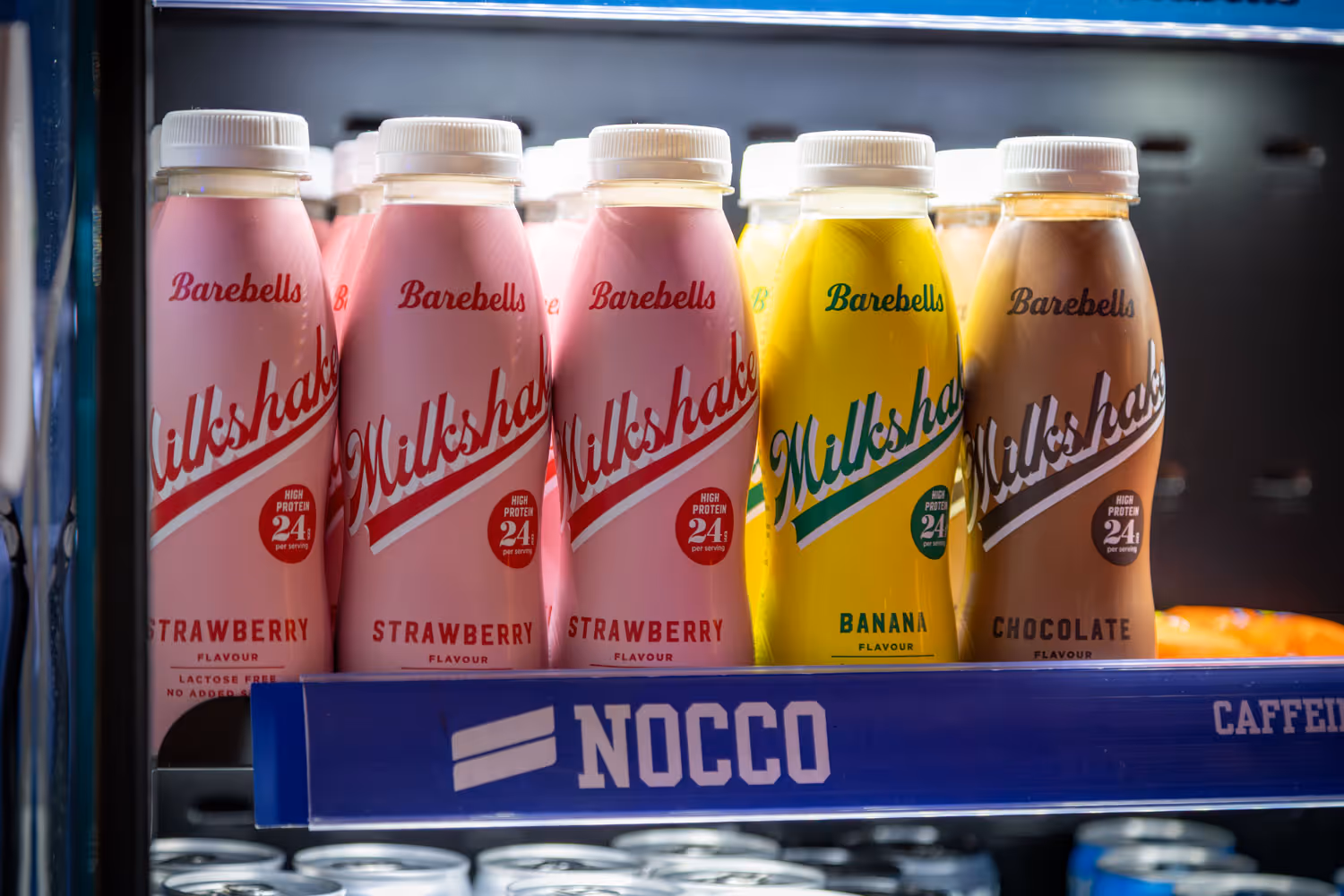 Bottles of Barebells high-protein milkshakes in strawberry, banana, and chocolate flavors on a store shelf.