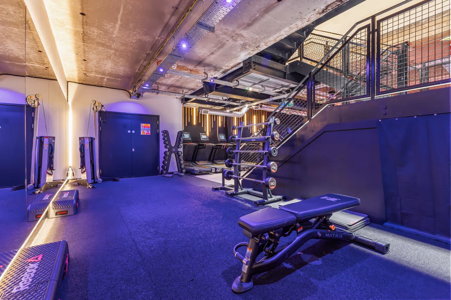 Modern gym area with weight bench, dumbbells, treadmills, step platforms, and lit ceiling accents.