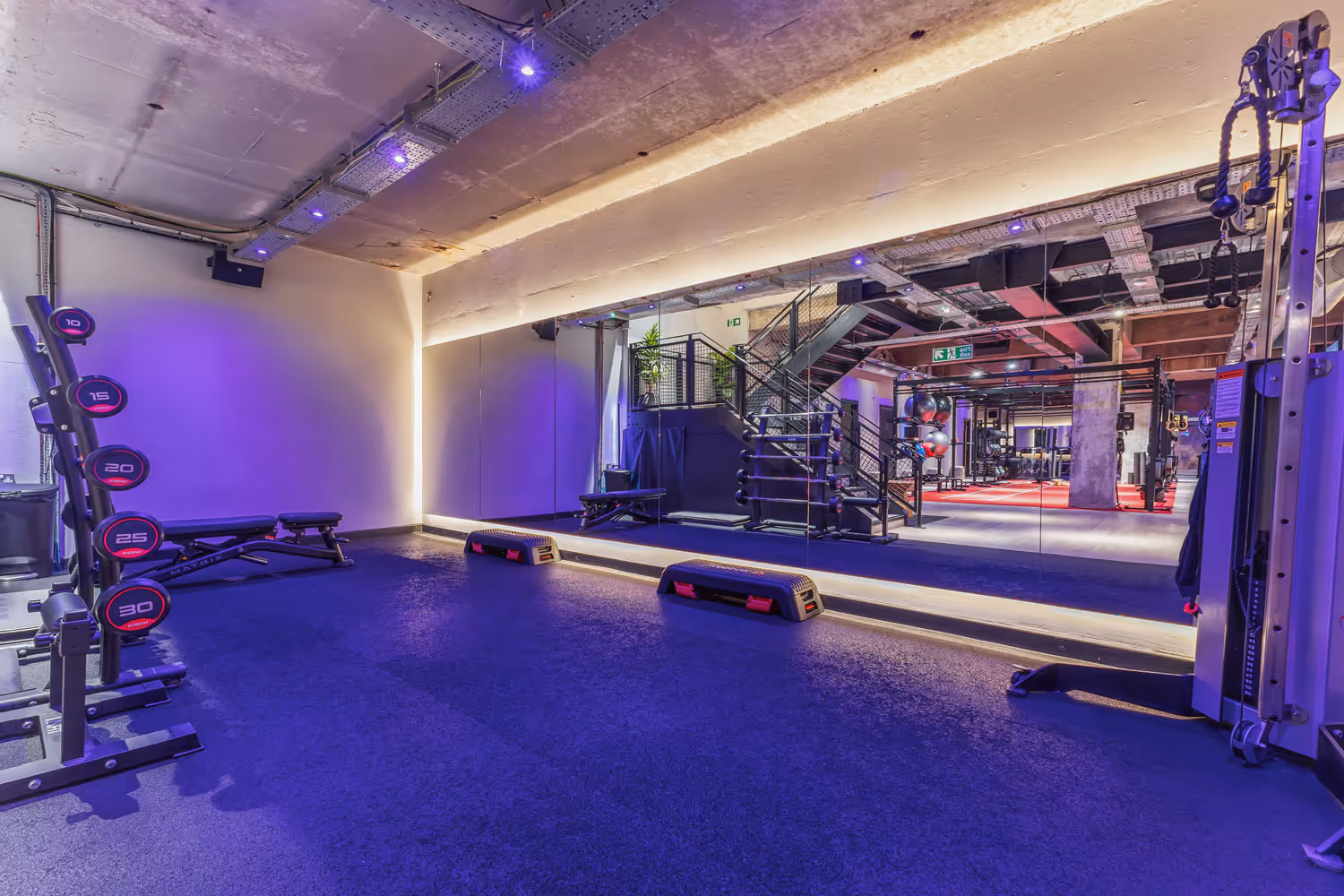 Modern gym area with weight rack, benches, step platforms, and mirrored wall under purple lighting.