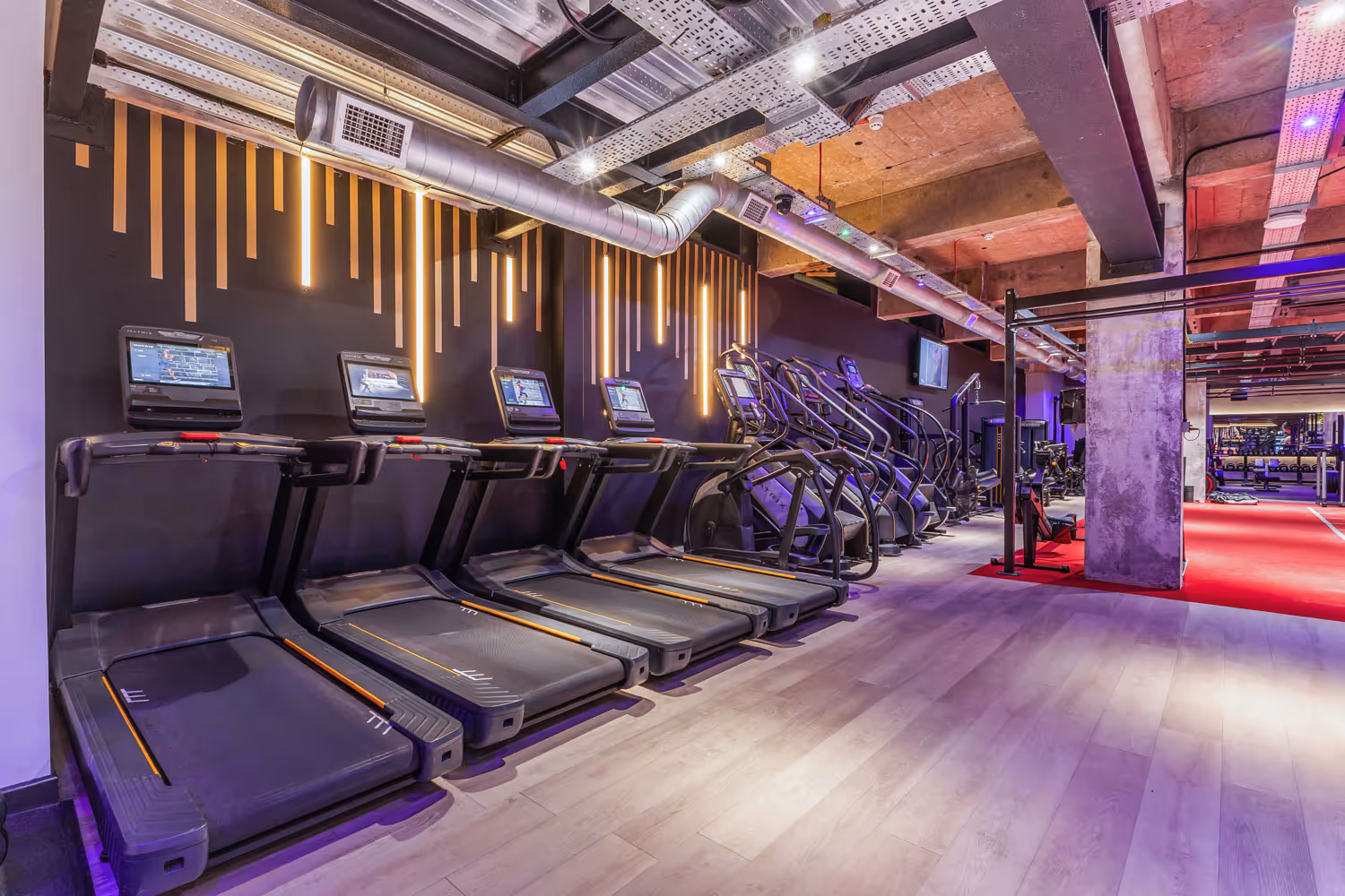 Row of treadmills and elliptical machines in a modern gym with decorative wall lights and exposed ceiling.