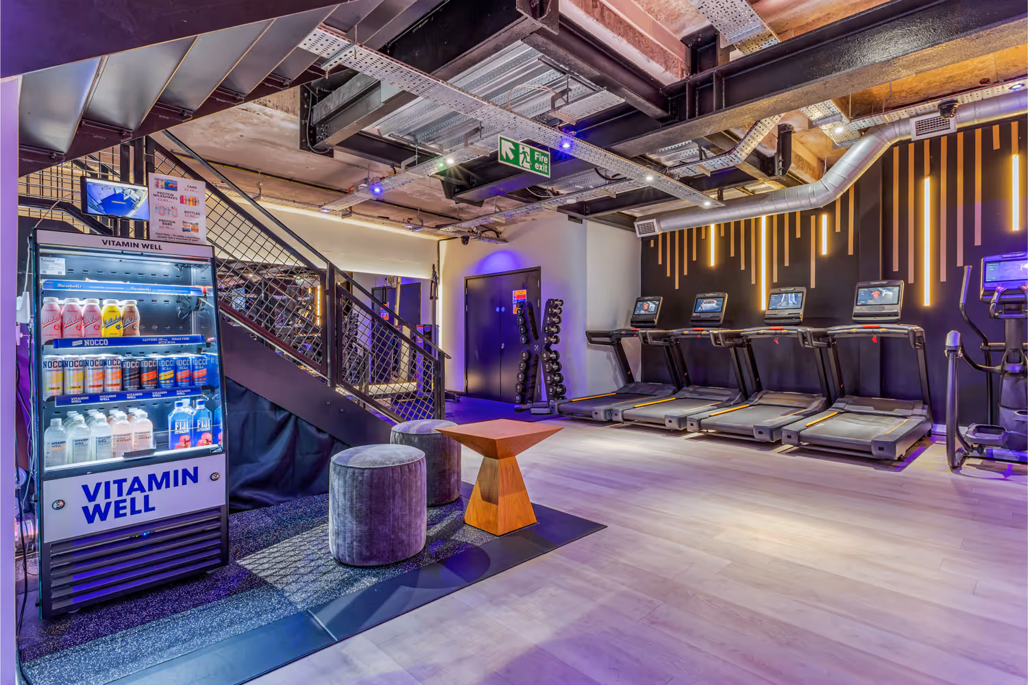Modern gym area with treadmills, dumbbells, Vitamin Well fridge, seating, and industrial ceiling.