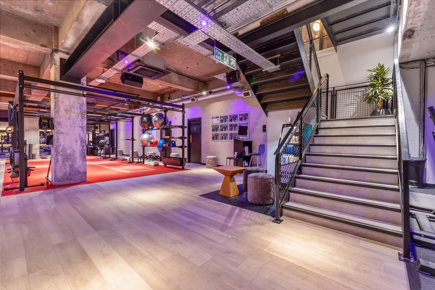 Modern gym interior with exercise equipment, red flooring area, stairs, and seating with a plant.
