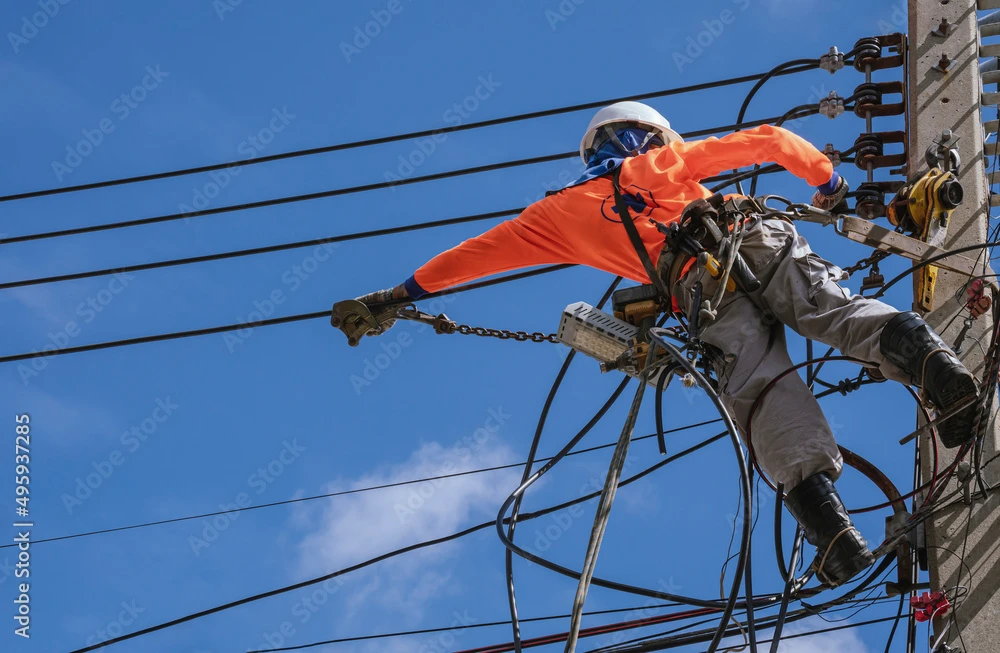 Utility worker in safety gear repairing power lines on a utility pole against a clear blue sky.