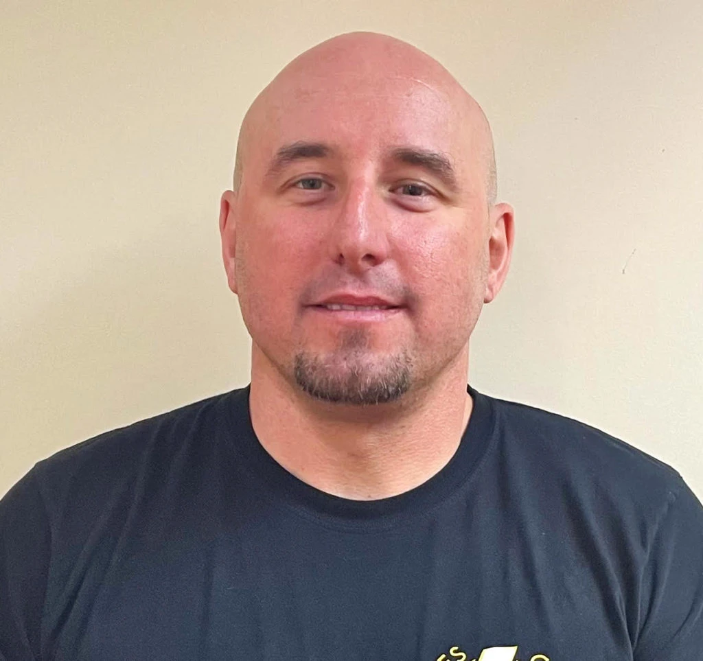 Man with shaved head, goatee, and black shirt standing against a plain beige wall.