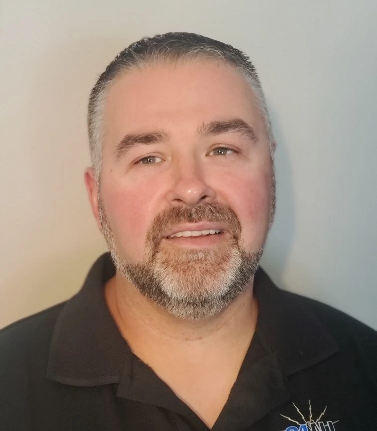Smiling middle-aged man with short gray-streaked hair and beard wearing a black collared shirt.