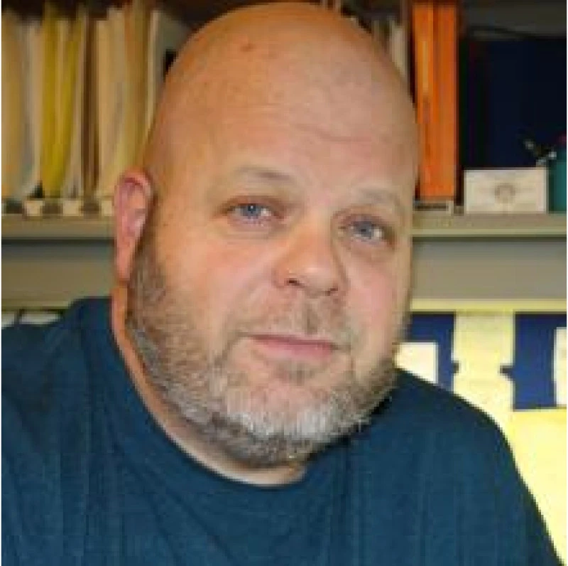 Bald man with a beard wearing a dark blue shirt, sitting indoors with shelves in the background.