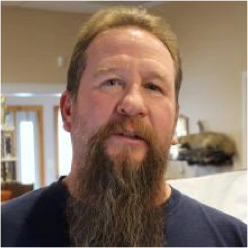 Man with a long beard and mustache wearing a dark shirt indoors.