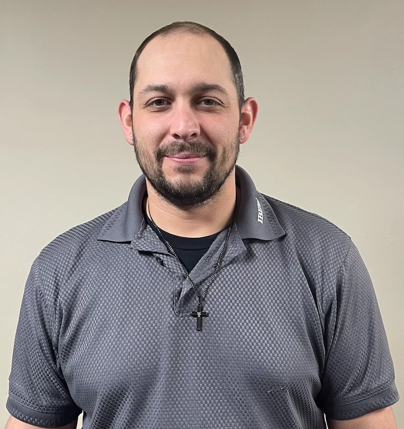 Man with short dark hair and beard wearing a gray patterned polo shirt and cross necklace.