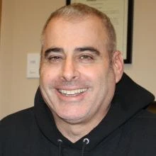 Smiling middle-aged man with short hair, wearing a black hoodie, indoors with a framed document behind.