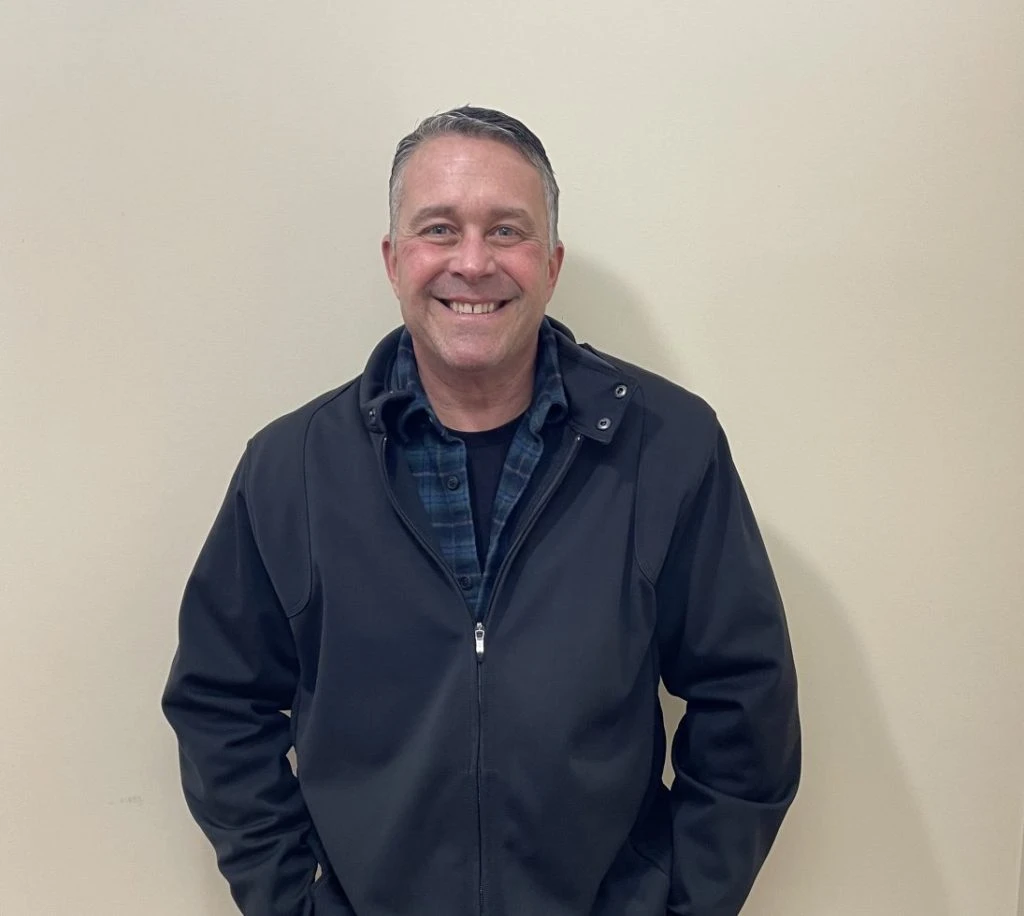 Smiling man wearing a dark jacket and plaid shirt standing against a plain beige wall.