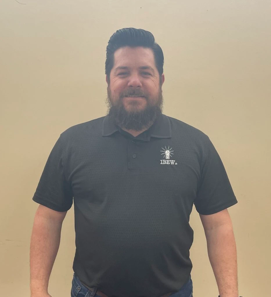 Man with beard and styled hair wearing a black IBEW polo shirt standing against a beige wall.