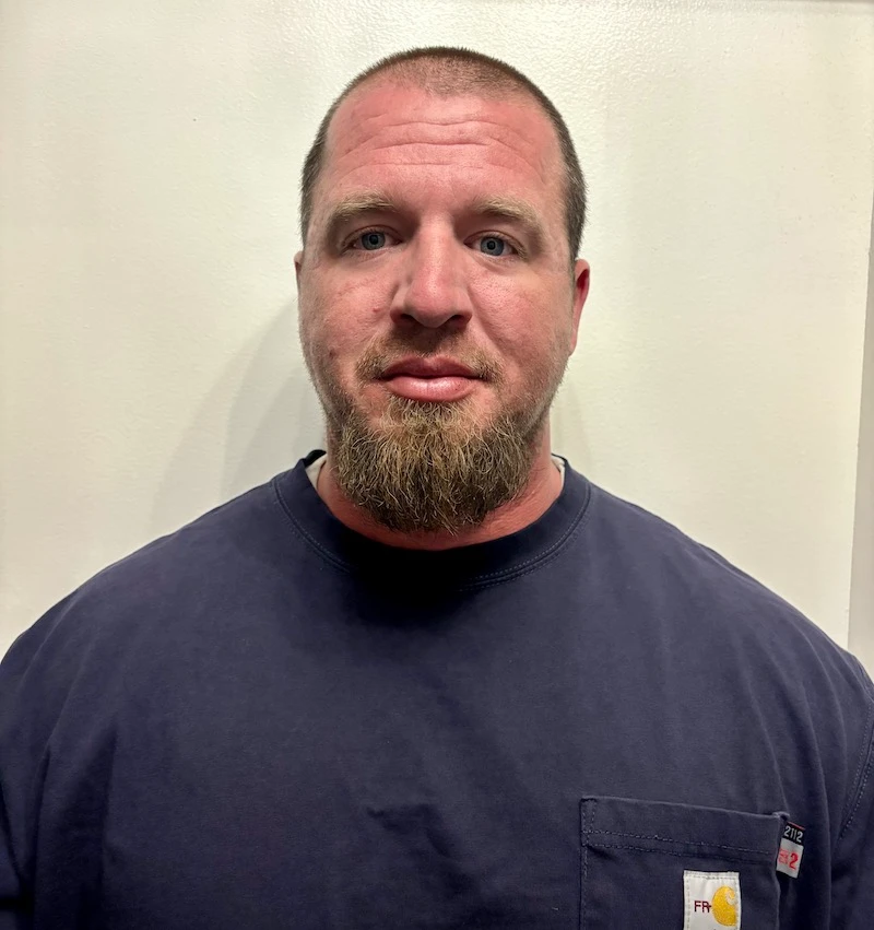 Man with short hair and beard wearing a dark blue shirt standing against a light wall.