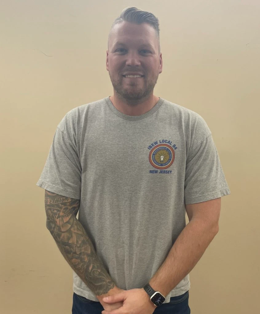 Smiling man with tattooed arm wearing a gray IBEW Local 94 New Jersey T-shirt and smartwatch.